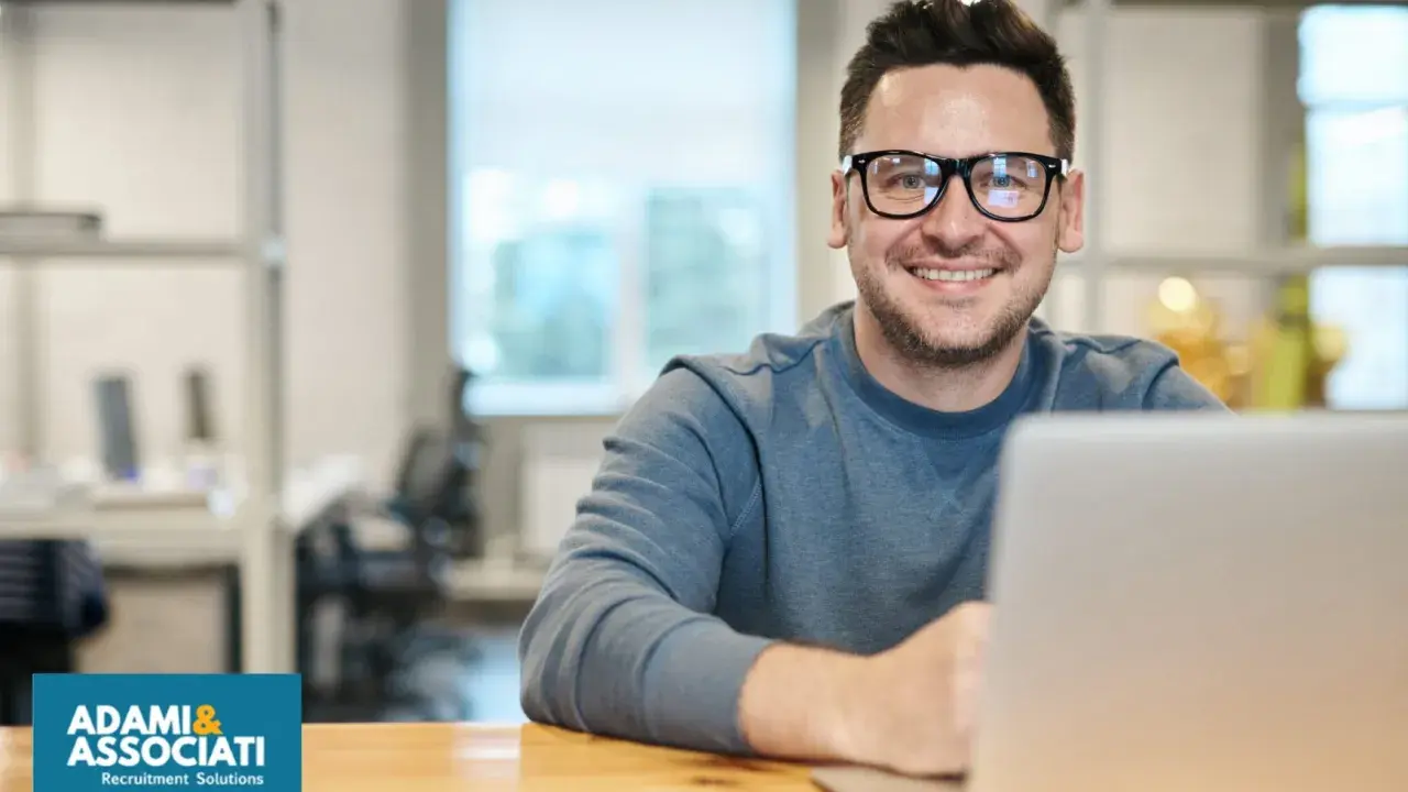 Uomo sorridente con occhiali, seduto alla scrivania con un laptop. Sembra un plant manager che gestisce il suo team.