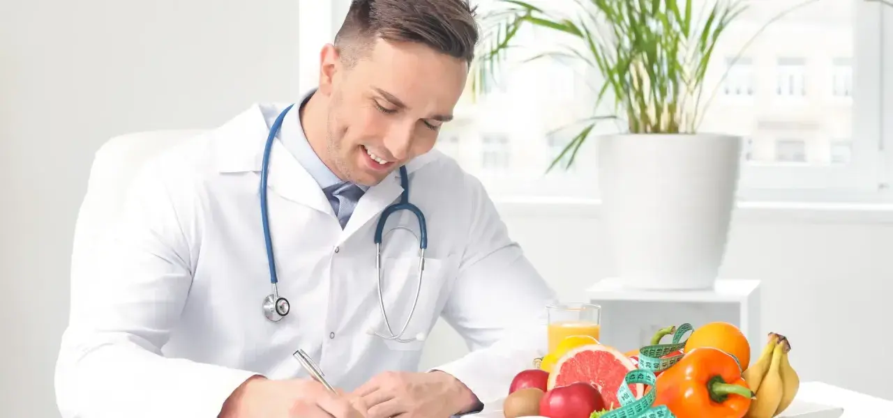 Medico sorridente con stetoscopio, circondato da frutta e verdura, mentre scrive. Un esempio di come diventare nutrizionista.