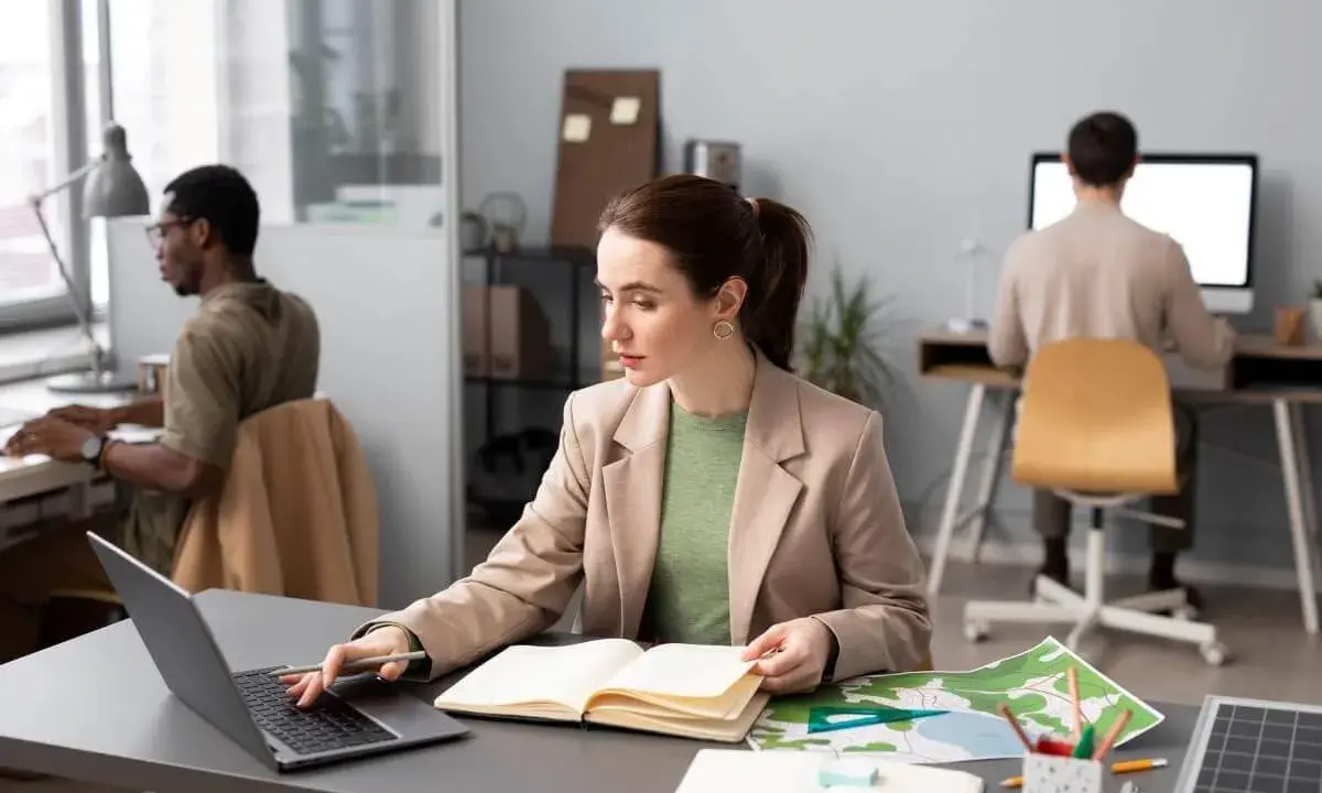 Donne al lavoro, forse preparano progetti per concorsi a Firenze. Uomini e donne concentrati sui loro computer, con mappe e quaderni aperti.
