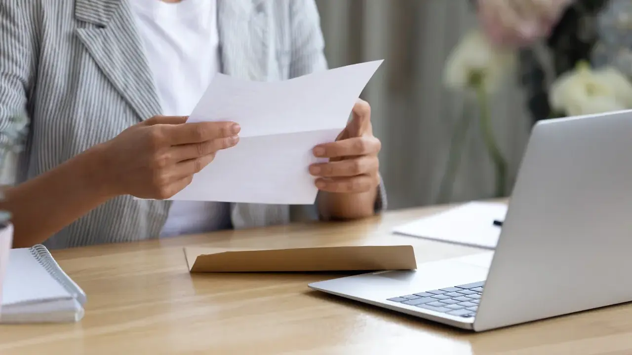 Persona esamina un documento, forse una busta paga contratto a chiamata, seduta alla scrivania con un laptop aperto.