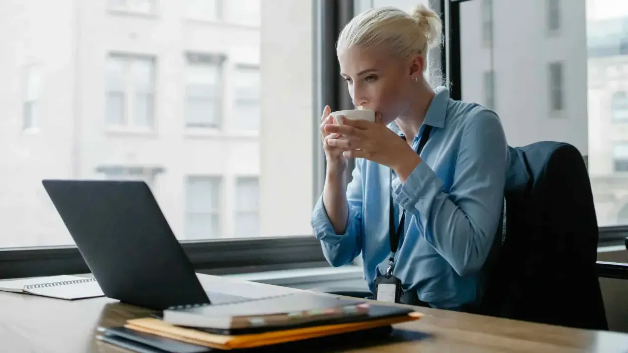 Donna bionda sorseggia caffè mentre lavora al laptop, cercando concorsi psicologi non scaduti.