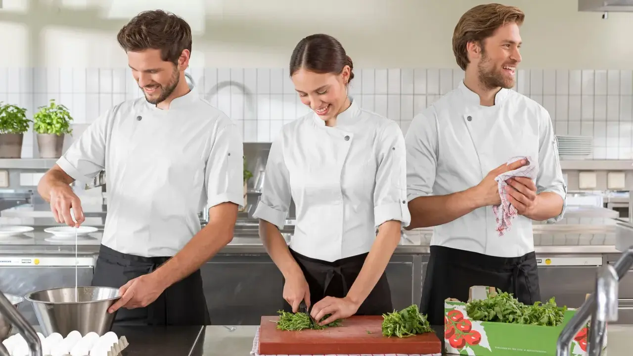 Tre commis di cucina, sorridenti, preparano ingredienti in una cucina professionale. Uno rompe un uovo, un'altra trita erbe aromatiche, il terzo asciuga le mani.