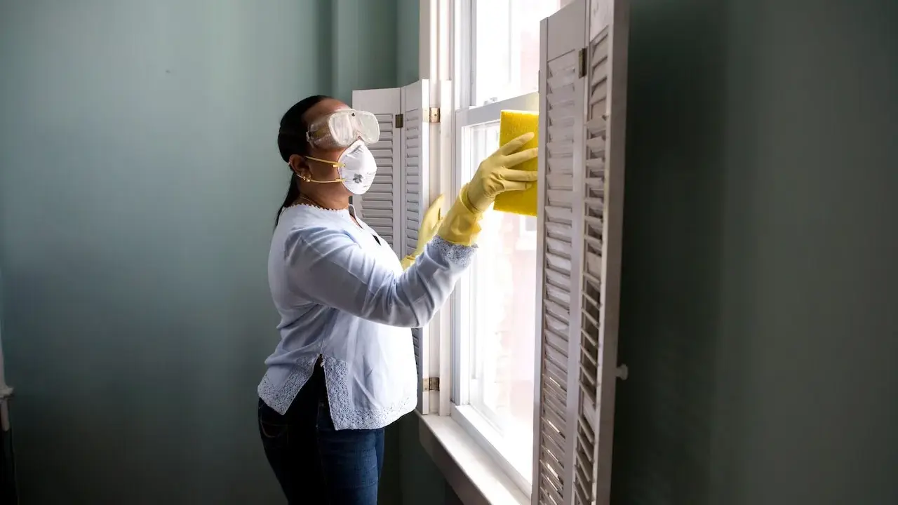 Donna con mascherina e occhiali pulisce una finestra con una spugna gialla. Il suo stipendio da addetta alle pulizie part time le permette di lavorare con cura.