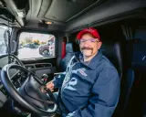 Un autista patente C sorride dal posto di guida del suo camion. Indossa una giacca blu con logo e un cappellino rosso.