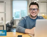 Uomo sorridente con occhiali, seduto alla scrivania con un laptop. Sembra un plant manager che gestisce il suo team.