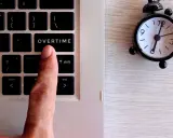 Dito, un dito preme il tasto "OVERTIME" su una tastiera, accanto a una sveglia. Quante ore di straordinario si possono fare in un mese?