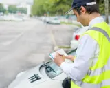 Un ausiliario del traffico con giubbotto catarifrangente scrive su un taccuino, controllando le auto parcheggiate.