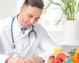 Medico sorridente con stetoscopio, circondato da frutta e verdura, mentre scrive. Un esempio di come diventare nutrizionista.
