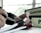 Mano che scrive su fogli con cartelline "Salaries" e "Pay" sullo sfondo, indicando il livello commercio.