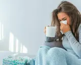 Donna con fazzoletto e tazza, coperta da un plaid. Il suo tirocinio è interrotto da una malattia.
