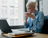 Donna bionda sorseggia caffè mentre lavora al laptop, cercando concorsi psicologi non scaduti.