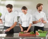 Tre commis di cucina, sorridenti, preparano ingredienti in una cucina professionale. Uno rompe un uovo, un'altra trita erbe aromatiche, il terzo asciuga le mani.