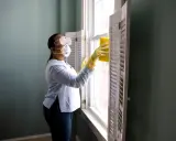 Donna con mascherina e occhiali pulisce una finestra con una spugna gialla. Il suo stipendio da addetta alle pulizie part time le permette di lavorare con cura.
