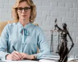 Donna con occhiali e camicia azzurra scrive a una scrivania, con la statua della giustizia sullo sfondo. Il percorso per diventare avvocato inizia qui.