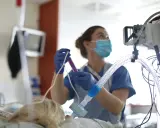 Un'infermiera con mascherina e guanti prepara una siringa, concentrata sul suo lavoro. Attrezzature mediche sullo sfondo.