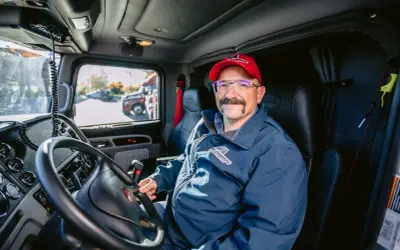 Un autista patente C sorride dal posto di guida del suo camion. Indossa una giacca blu con logo e un cappellino rosso.