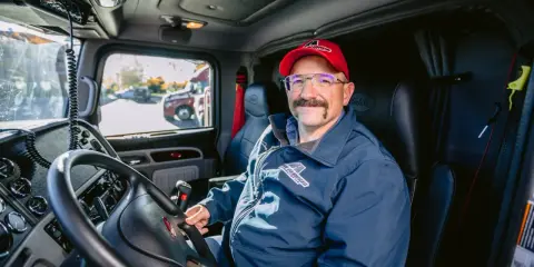 Un autista patente C sorride dal posto di guida del suo camion. Indossa una giacca blu con logo e un cappellino rosso.