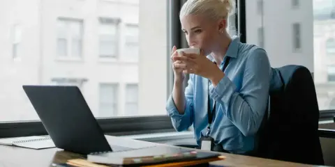 Donna bionda sorseggia caffè mentre lavora al laptop, cercando concorsi psicologi non scaduti.