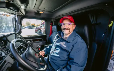 Un autista patente C sorride dal posto di guida del suo camion. Indossa una giacca blu con logo e un cappellino rosso.