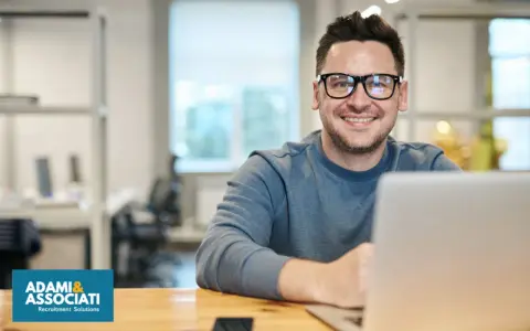 Uomo sorridente con occhiali, seduto alla scrivania con un laptop. Sembra un plant manager che gestisce il suo team.