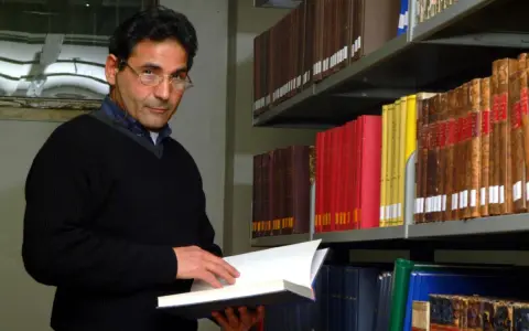Uomo con occhiali in biblioteca, sfoglia un libro. Forse pensa al suo bibliotecario stipendio.