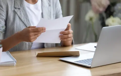 Persona esamina un documento, forse una busta paga contratto a chiamata, seduta alla scrivania con un laptop aperto.