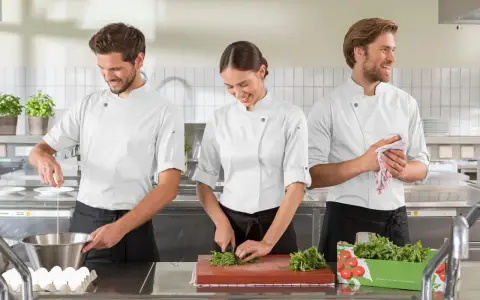 Tre commis di cucina, sorridenti, preparano ingredienti in una cucina professionale. Uno rompe un uovo, un'altra trita erbe aromatiche, il terzo asciuga le mani.