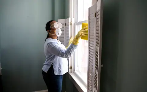 Donna con mascherina e occhiali pulisce una finestra con una spugna gialla. Il suo stipendio da addetta alle pulizie part time le permette di lavorare con cura.