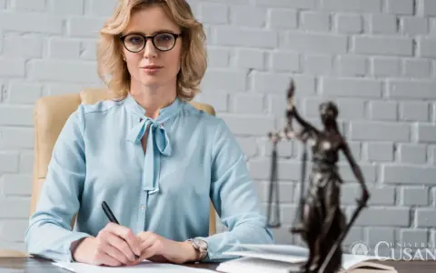 Donna con occhiali e camicia azzurra scrive a una scrivania, con la statua della giustizia sullo sfondo. Il percorso per diventare avvocato inizia qui.
