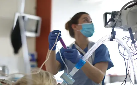 Un'infermiera con mascherina e guanti prepara una siringa, concentrata sul suo lavoro. Attrezzature mediche sullo sfondo.