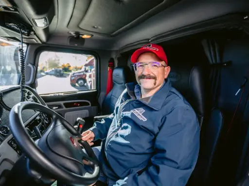 Un autista patente C sorride dal posto di guida del suo camion. Indossa una giacca blu con logo e un cappellino rosso.