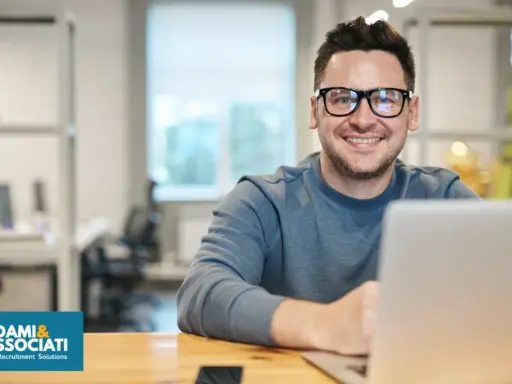 Uomo sorridente con occhiali, seduto alla scrivania con un laptop. Sembra un plant manager che gestisce il suo team.