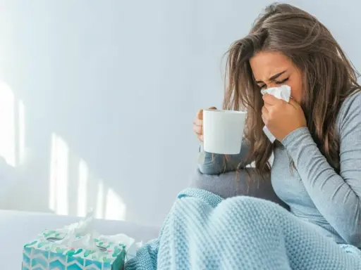 Donna con fazzoletto e tazza, coperta da un plaid. Il suo tirocinio è interrotto da una malattia.