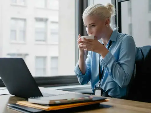 Donna bionda sorseggia caffè mentre lavora al laptop, cercando concorsi psicologi non scaduti.
