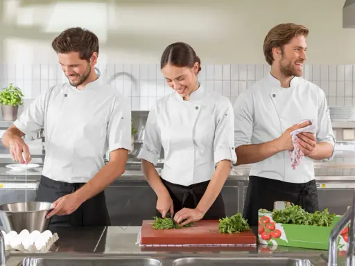 Tre commis di cucina, sorridenti, preparano ingredienti in una cucina professionale. Uno rompe un uovo, un'altra trita erbe aromatiche, il terzo asciuga le mani.