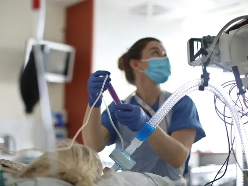 Un'infermiera con mascherina e guanti prepara una siringa, concentrata sul suo lavoro. Attrezzature mediche sullo sfondo.