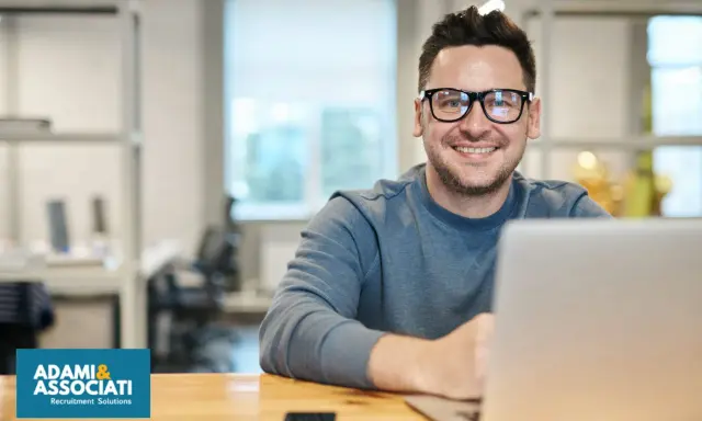 Uomo sorridente con occhiali, seduto alla scrivania con un laptop. Sembra un plant manager che gestisce il suo team.