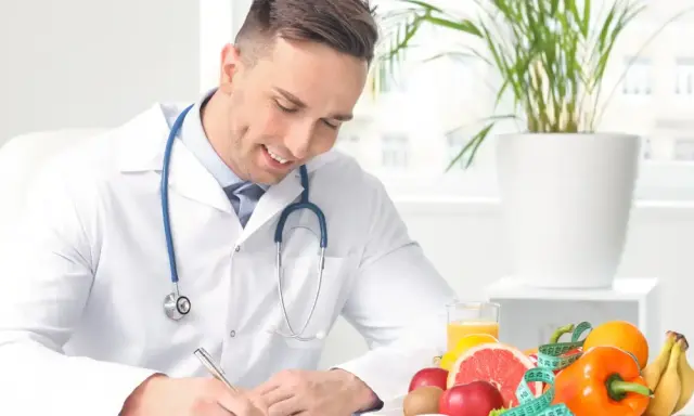 Medico sorridente con stetoscopio, circondato da frutta e verdura, mentre scrive. Un esempio di come diventare nutrizionista.