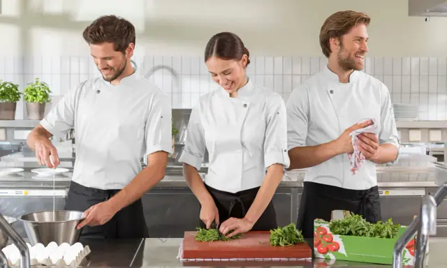 Tre commis di cucina, sorridenti, preparano ingredienti in una cucina professionale. Uno rompe un uovo, un'altra trita erbe aromatiche, il terzo asciuga le mani.