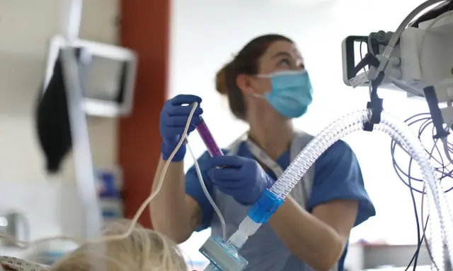 Un'infermiera con mascherina e guanti prepara una siringa, concentrata sul suo lavoro. Attrezzature mediche sullo sfondo.