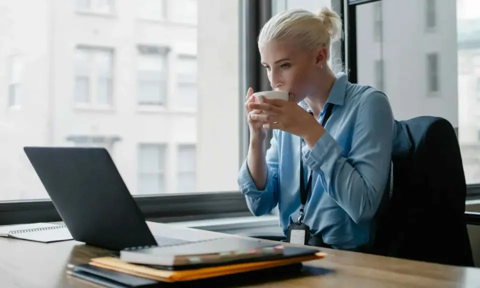 Donna bionda sorseggia caffè mentre lavora al laptop, cercando concorsi psicologi non scaduti.