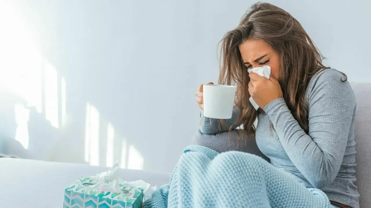 Donna con fazzoletto e tazza, coperta da un plaid. Il suo tirocinio è interrotto da una malattia.