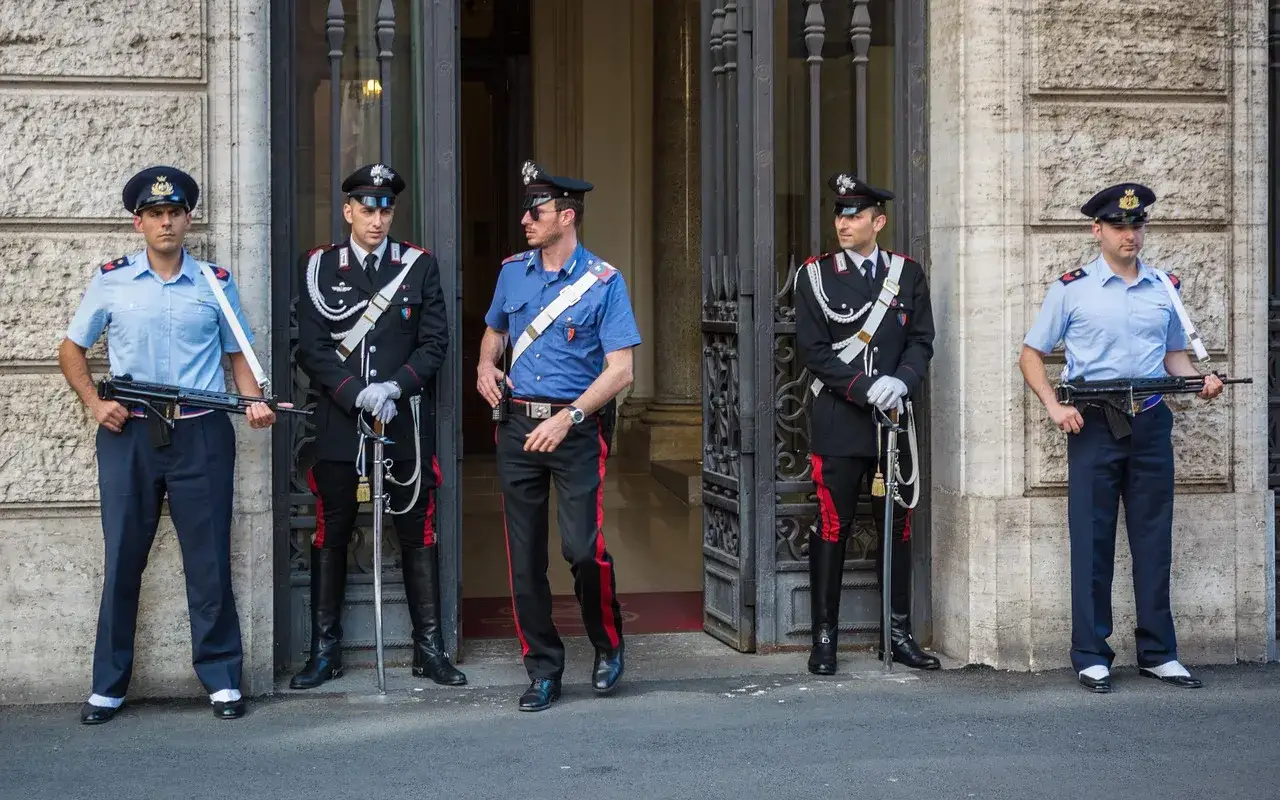 Carabinieri in divisa, alcuni con fucile, altri con sciabola, sorvegliano un ingresso. Il loro stipendio è meritato per la dedizione.