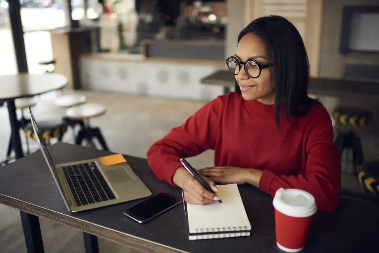 Donna con occhiali scrive su un quaderno, laptop e caff&egrave; accanto. Un'ispirazione per il suo lavoro di copywriter.