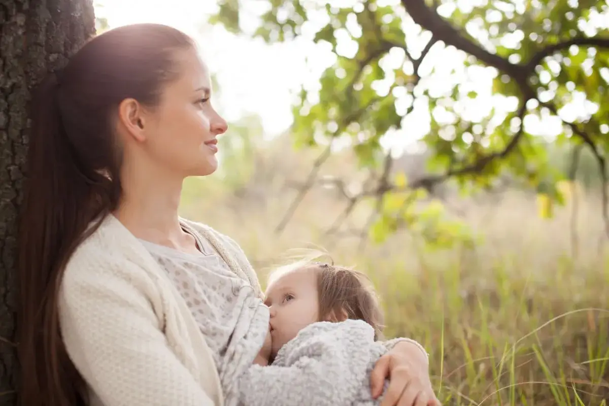 Mamma appoggiata a un albero allatta il suo bambino, un momento di tenerezza che celebra il permesso allattamento nella natura.