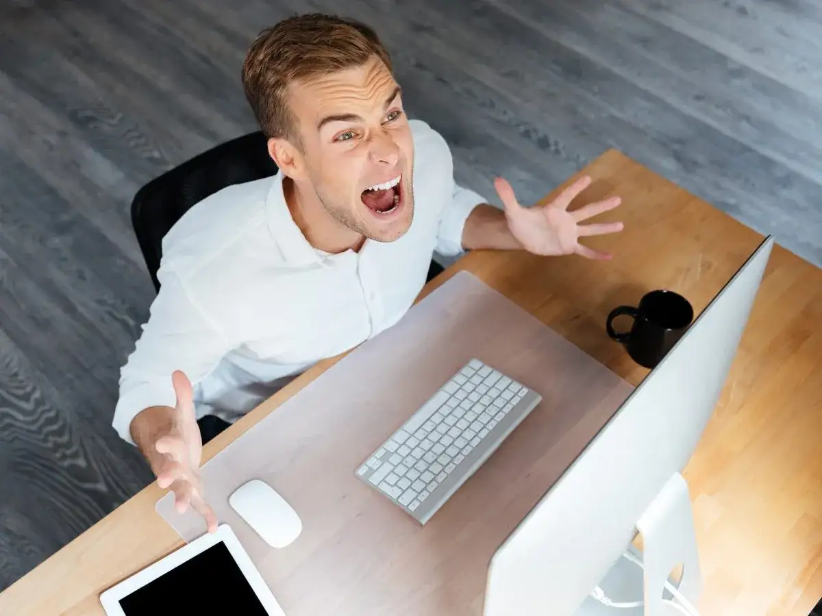 Uomo in camicia bianca urla davanti al computer, mani aperte in segno di frustrazione. Un chiaro segnale di un ambiente lavoro tossico.