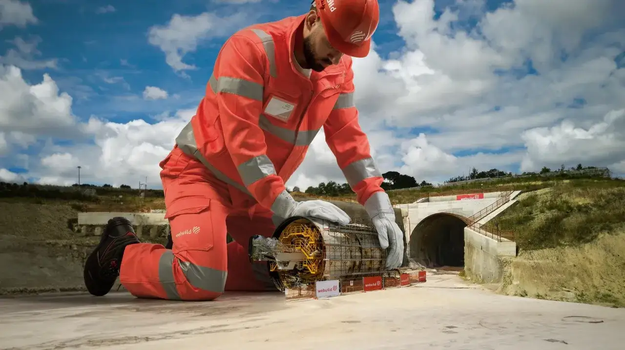 Un geometra di cantiere in tuta arancione esamina un macchinario per scavi. Sullo sfondo, l'imbocco di un tunnel.