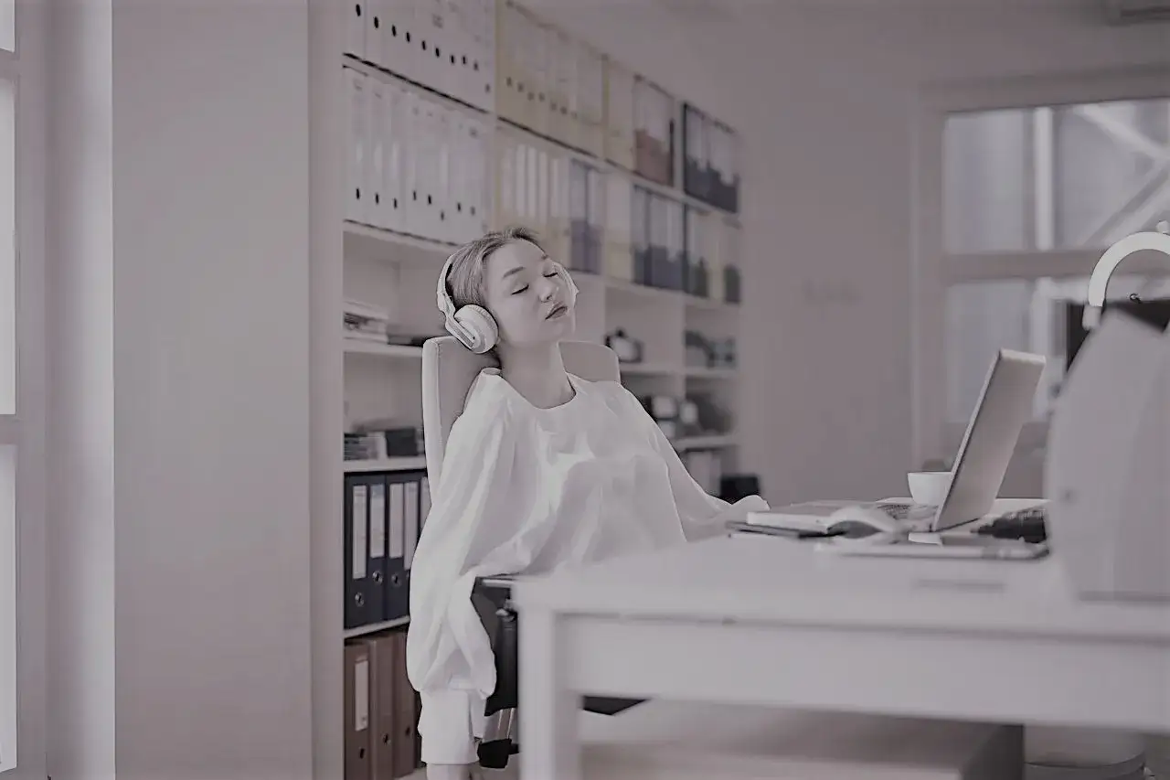 Donna con cuffie, occhi chiusi, in ufficio. Un esempio di quiet quitting, che si prende una pausa mentale dal lavoro.