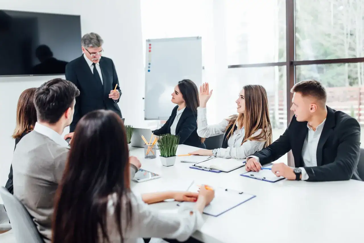 Un uomo in giacca e cravatta parla a un gruppo di giovani professionisti durante un one to one meeting. Una ragazza alza la mano per fare una domanda.