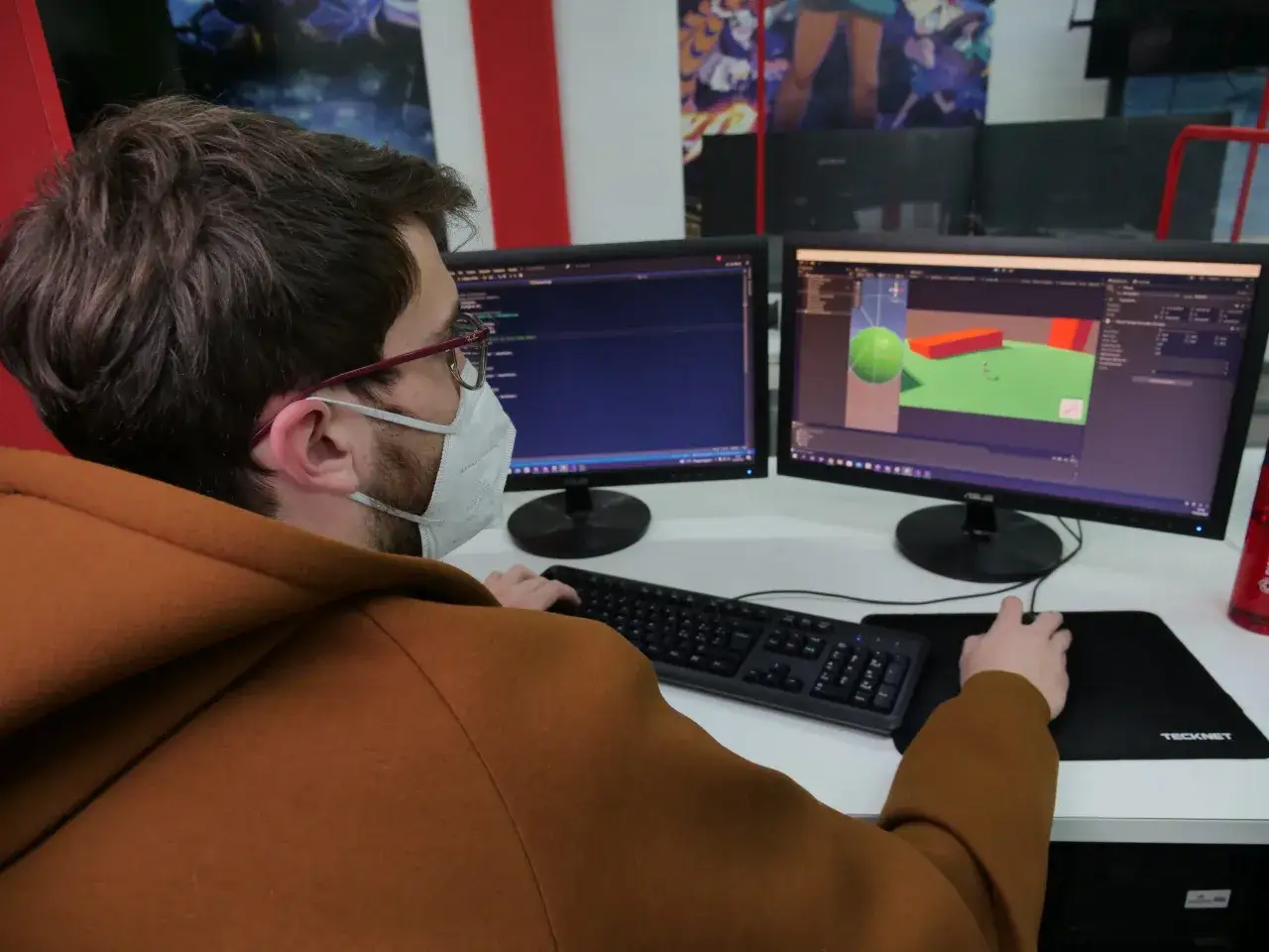 Un programmatore con mascherina lavora su due monitor, uno mostra codice, l'altro un ambiente 3D. Chiss&agrave; quanto guadagna un programmatore cos&igrave; concentrato!